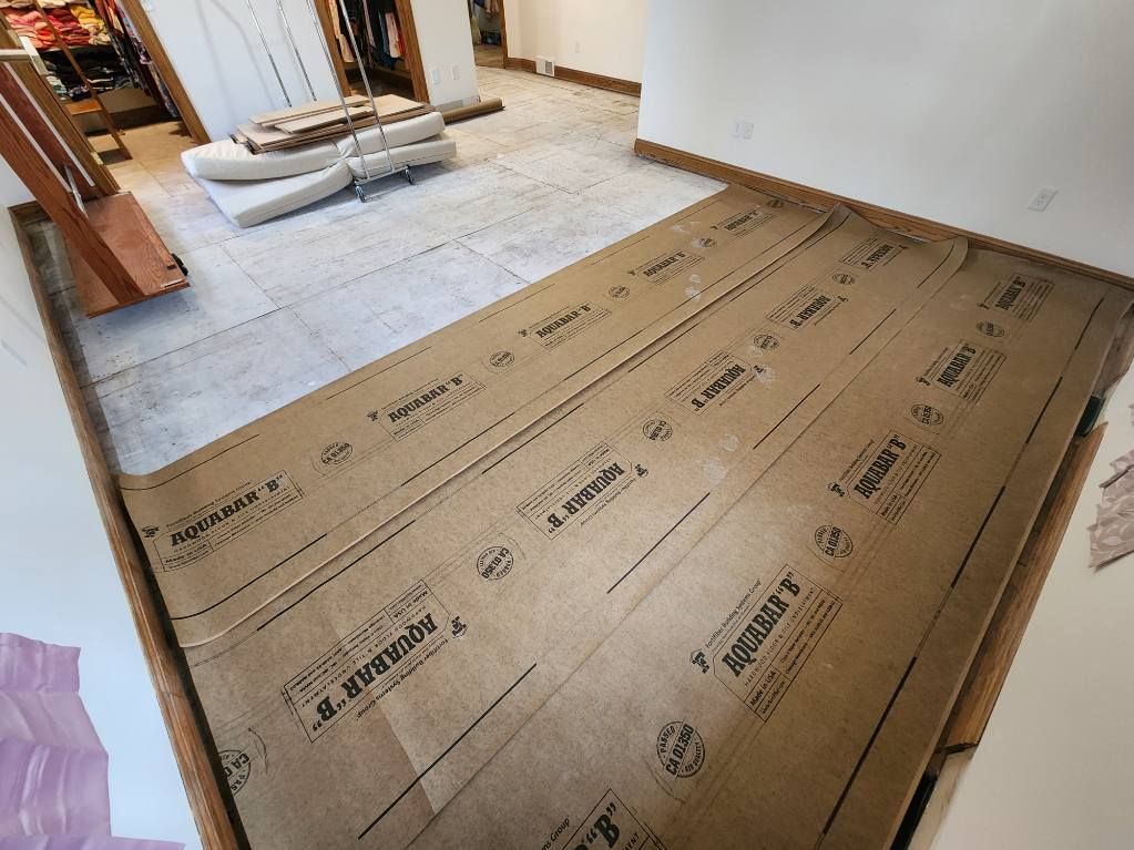 Flooring underlayment installation in a room; brown paper rolls, white foam, and wooden planks visible.