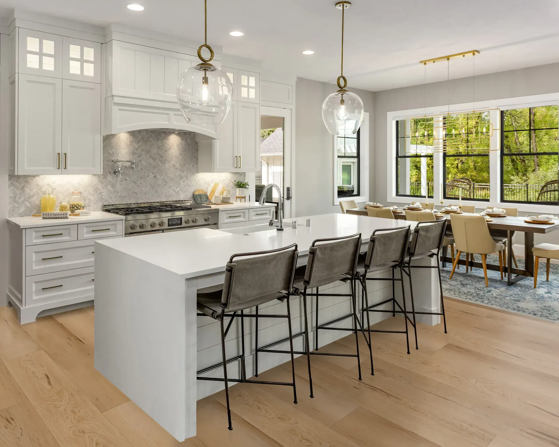 Bright white kitchen with island and seating, connected to dining area with large windows overlooking trees.