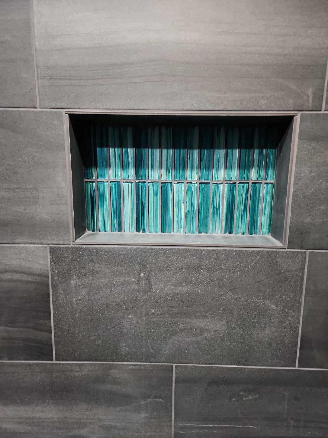 Recessed niche in dark gray tile filled with vertical turquoise glass strips.