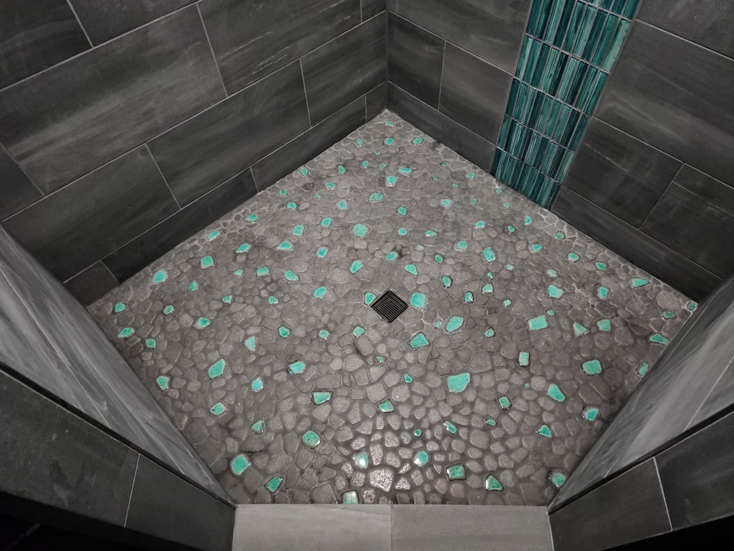 Shower with dark gray tiled walls and floor of textured gray pebbles with turquoise accents.