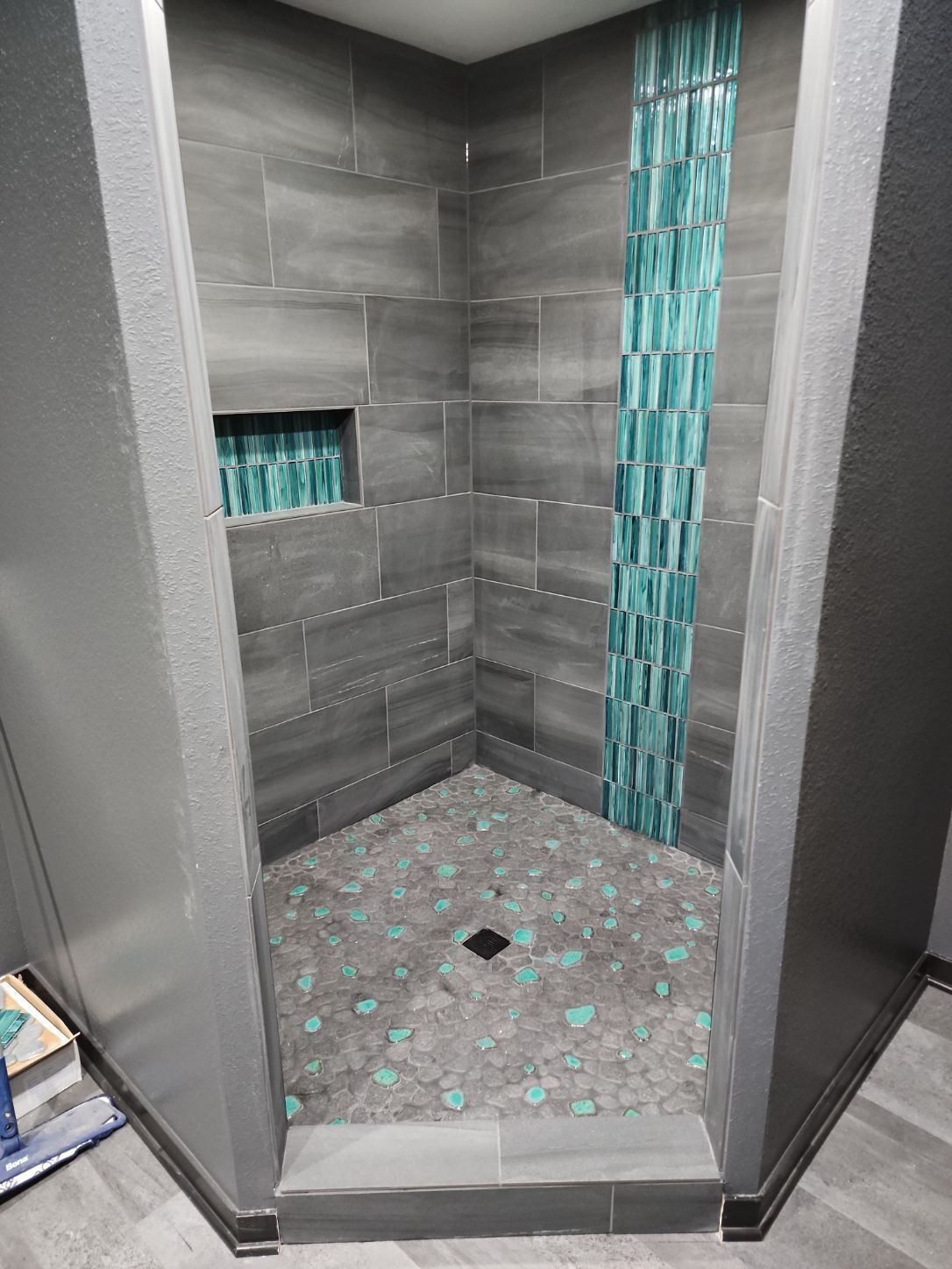 Gray tiled shower with turquoise accent strip and pebble floor.