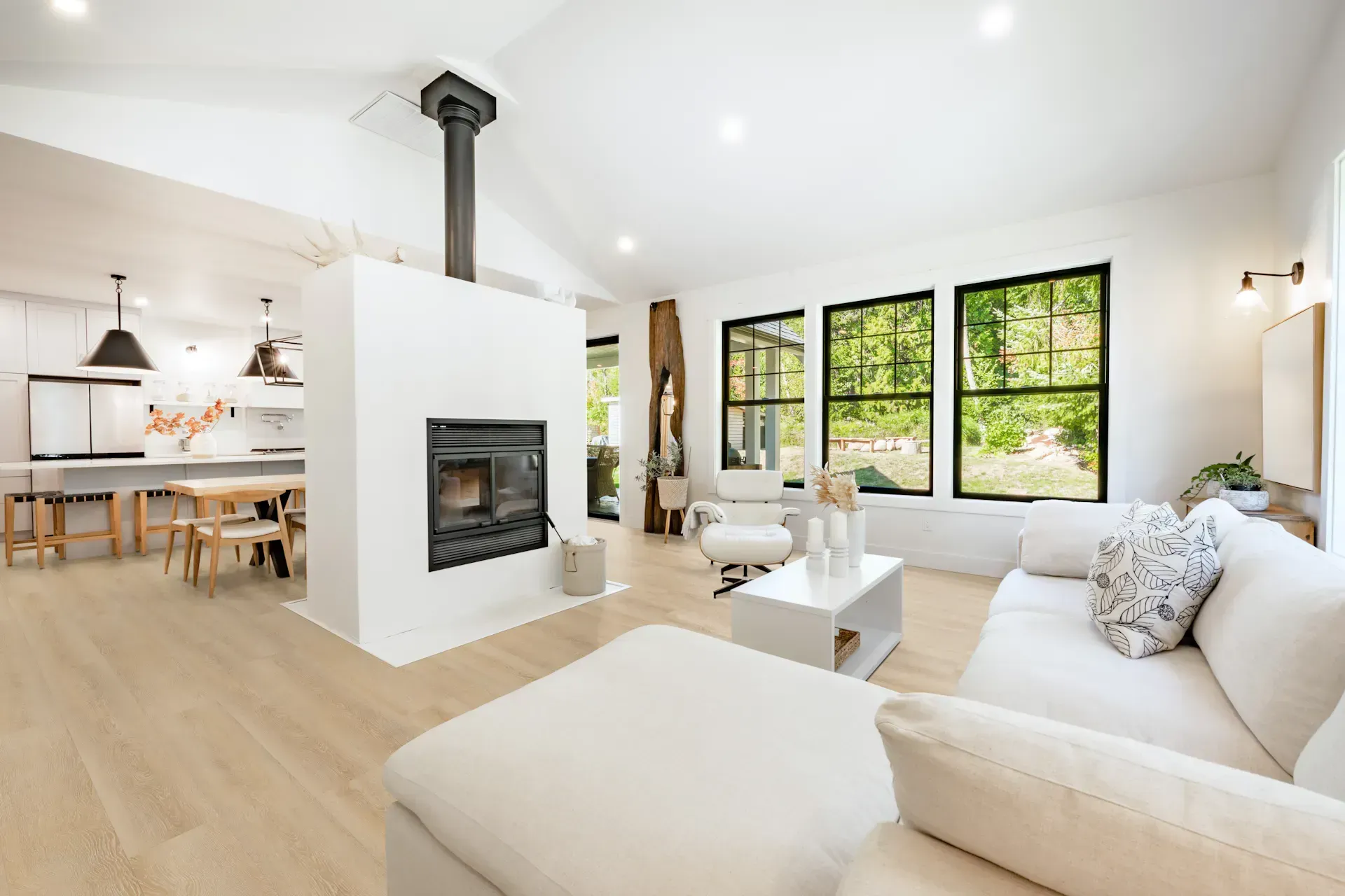 Bright, modern living room with fireplace, windows, and white furniture; open to kitchen.