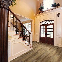 A grand foyer with stairs, double doors, and wood-look flooring. Beige walls and dark brown accents.