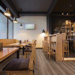 Interior of a restaurant with wooden tables, chairs, and bar.  Wine bottles on shelves and a light wood floor.