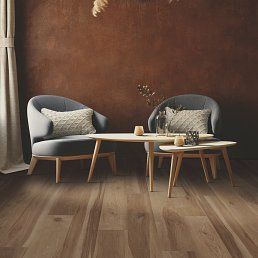 Two gray armchairs and wood tables in a room with brown walls and wood floors.