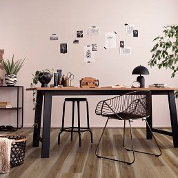 Desk in a room with a black wire chair, stool, lamp, and a plant against a light pink wall.