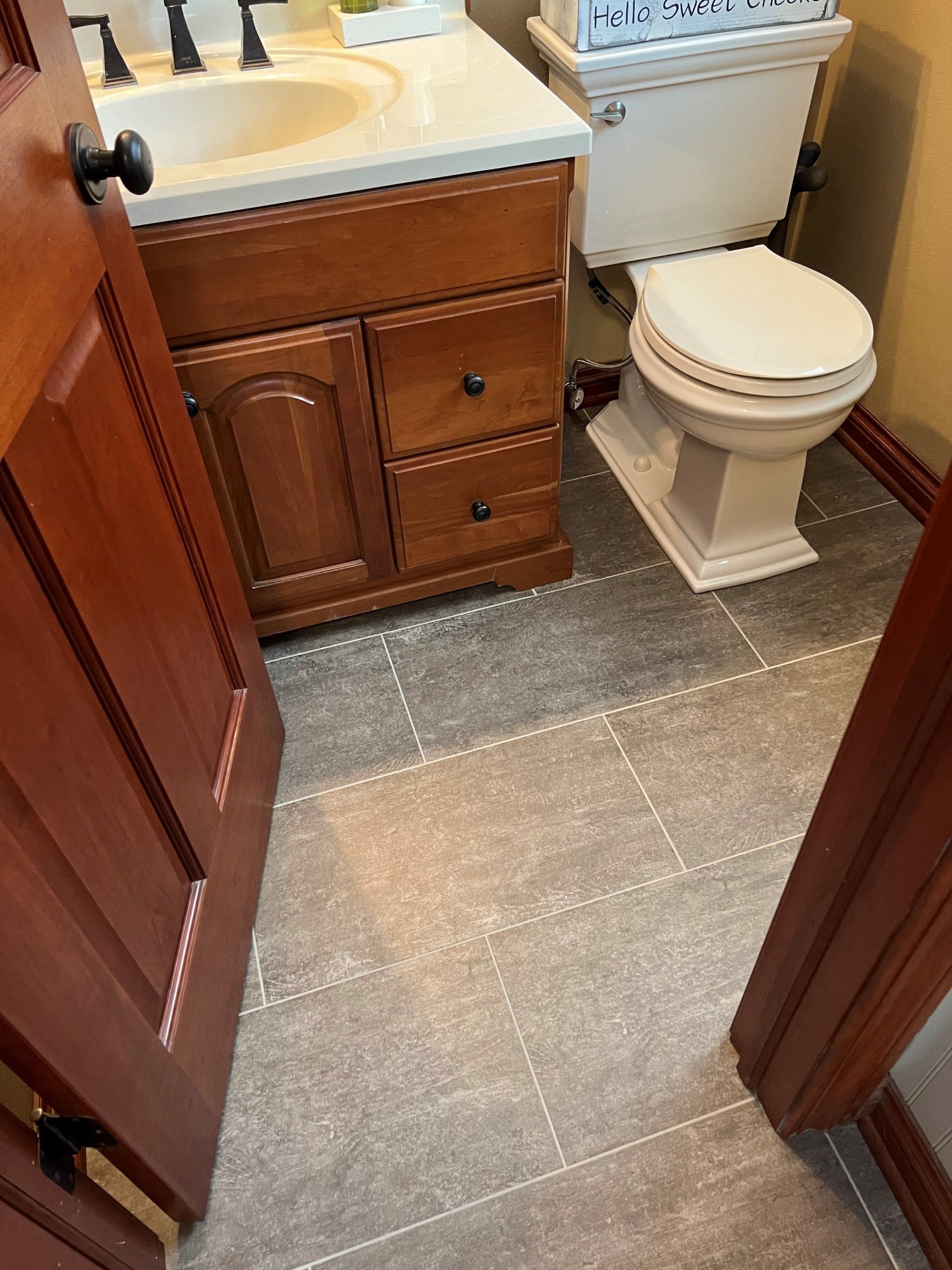 Bathroom with wooden vanity, toilet, and gray tile flooring. Partially open brown door on the left.