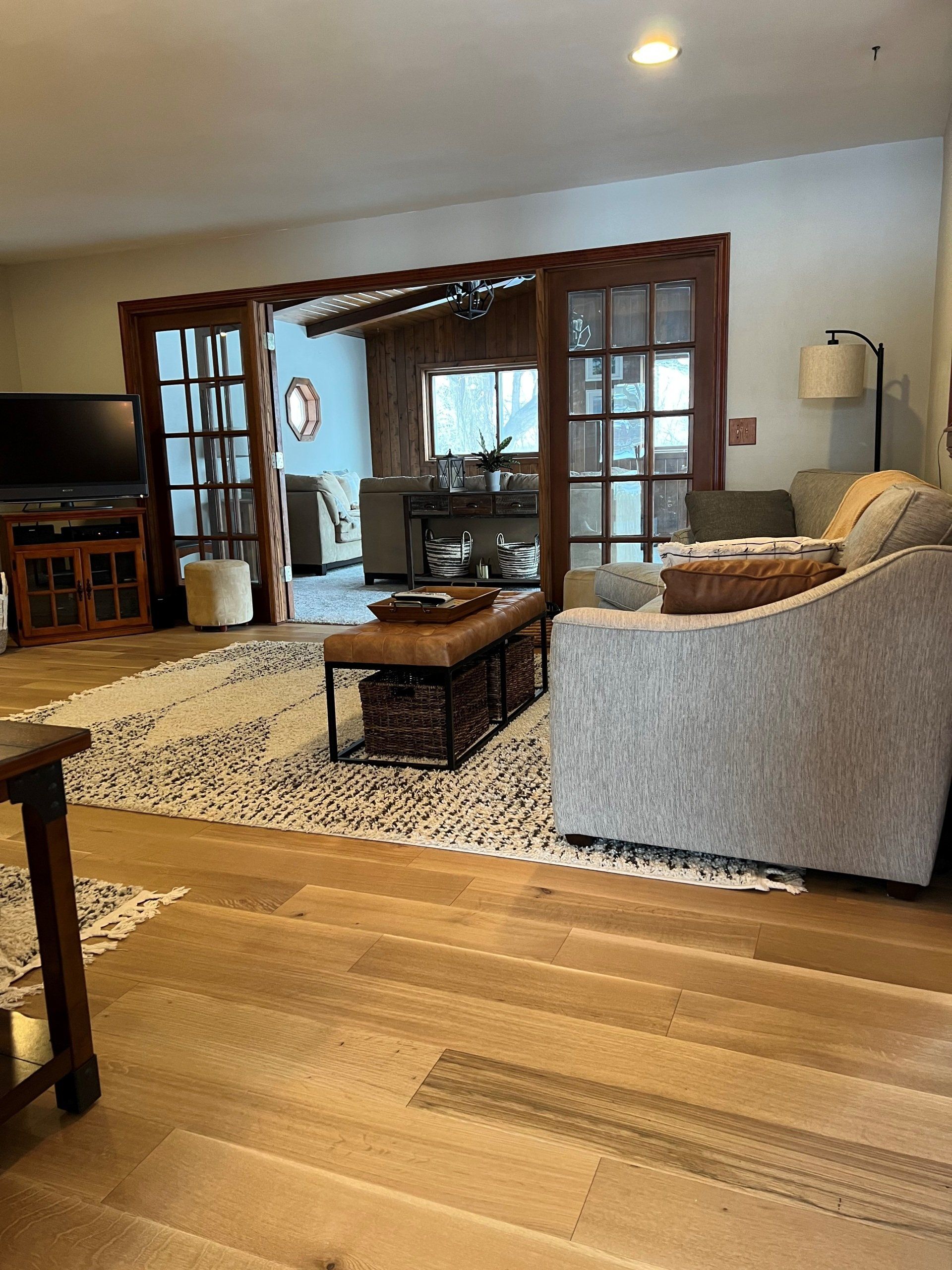 Living room with wood floors, rug, grey couch, and brown French doors leading to another room.