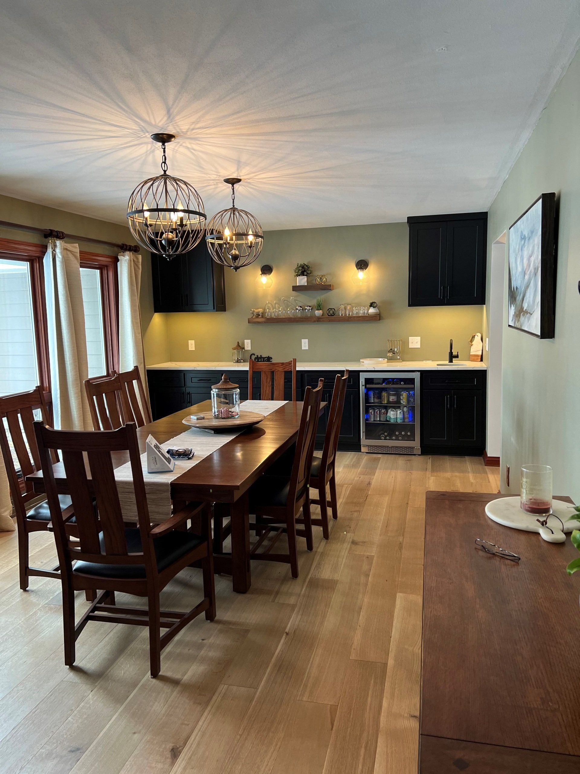 Dining room with a long wooden table, black cabinets, and two chandelier lights.