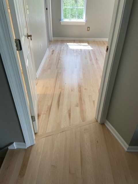 Light wood flooring in a hallway leading into a bedroom with a window; pale walls and white trim.