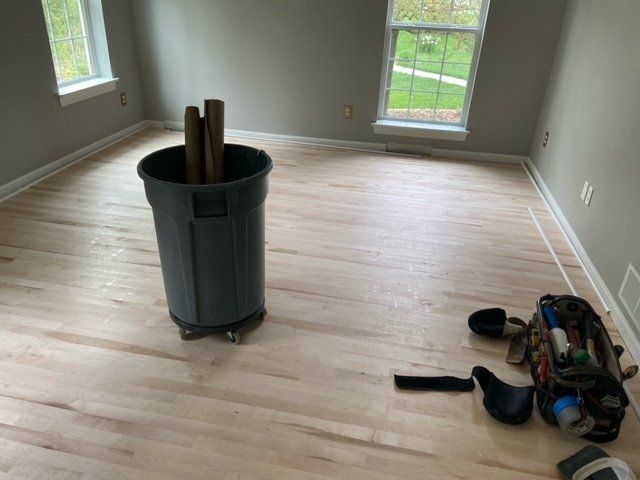 Room with refinished hardwood floor, gray walls, trash can, and tools; windows on both sides.