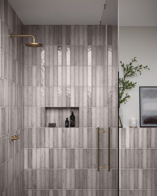 Shower with vertical gray tiles, a brass shower head, glass door, and built-in niche with toiletries.