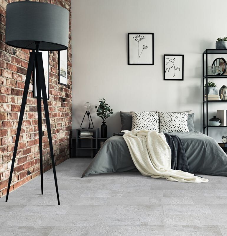 Bedroom with brick wall, gray bed, floor lamp, and black framed art.