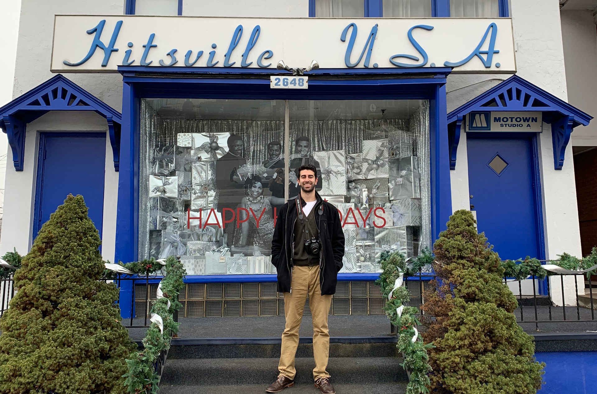 Alex Macksoud standing in front of Hitsville U.S.A. building with blue trim, windows decorated with tinsel, and evergreen bushes.