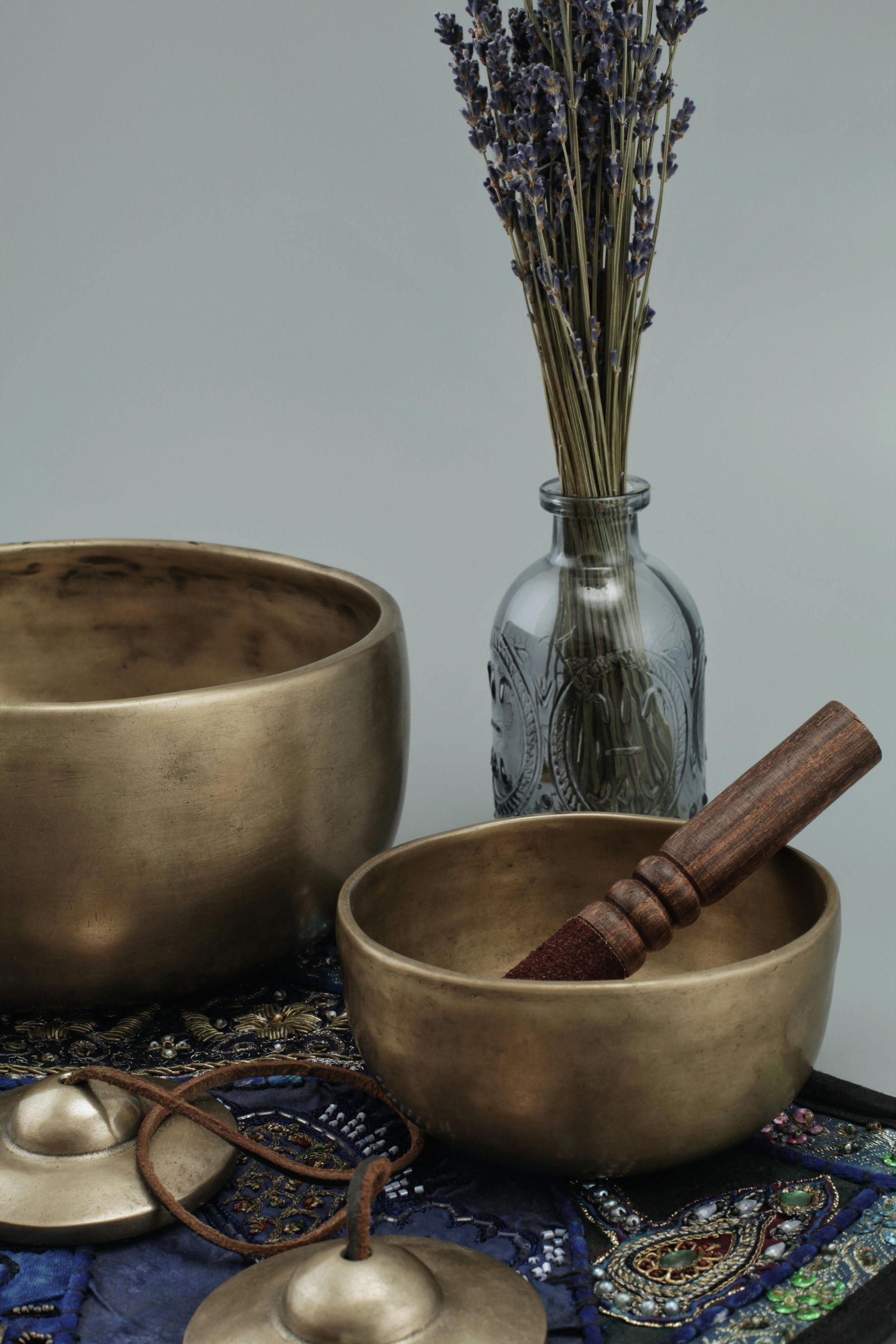 Two bronze singing bowls, cymbals, lavender in vase, on patterned mat.