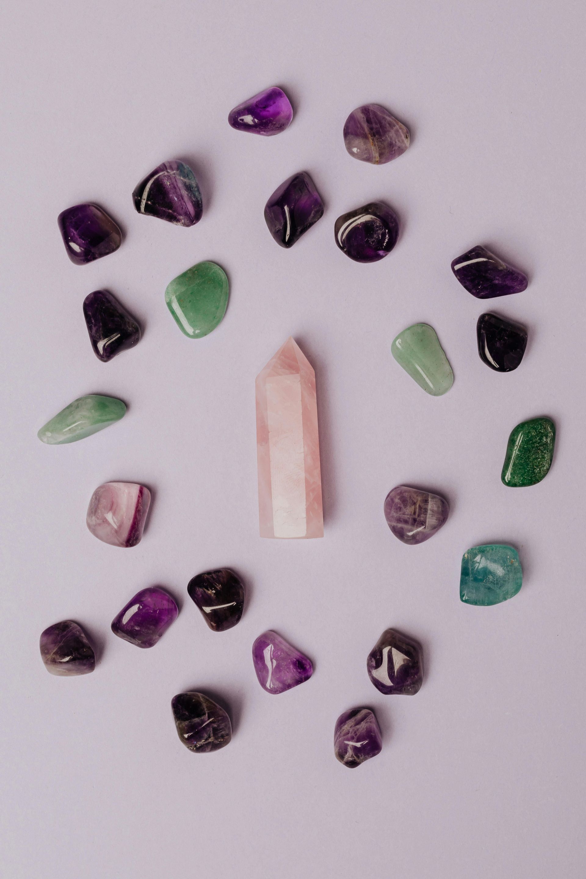 A collection of amethyst and green stones arranged around a central rose quartz crystal on a lavender surface.