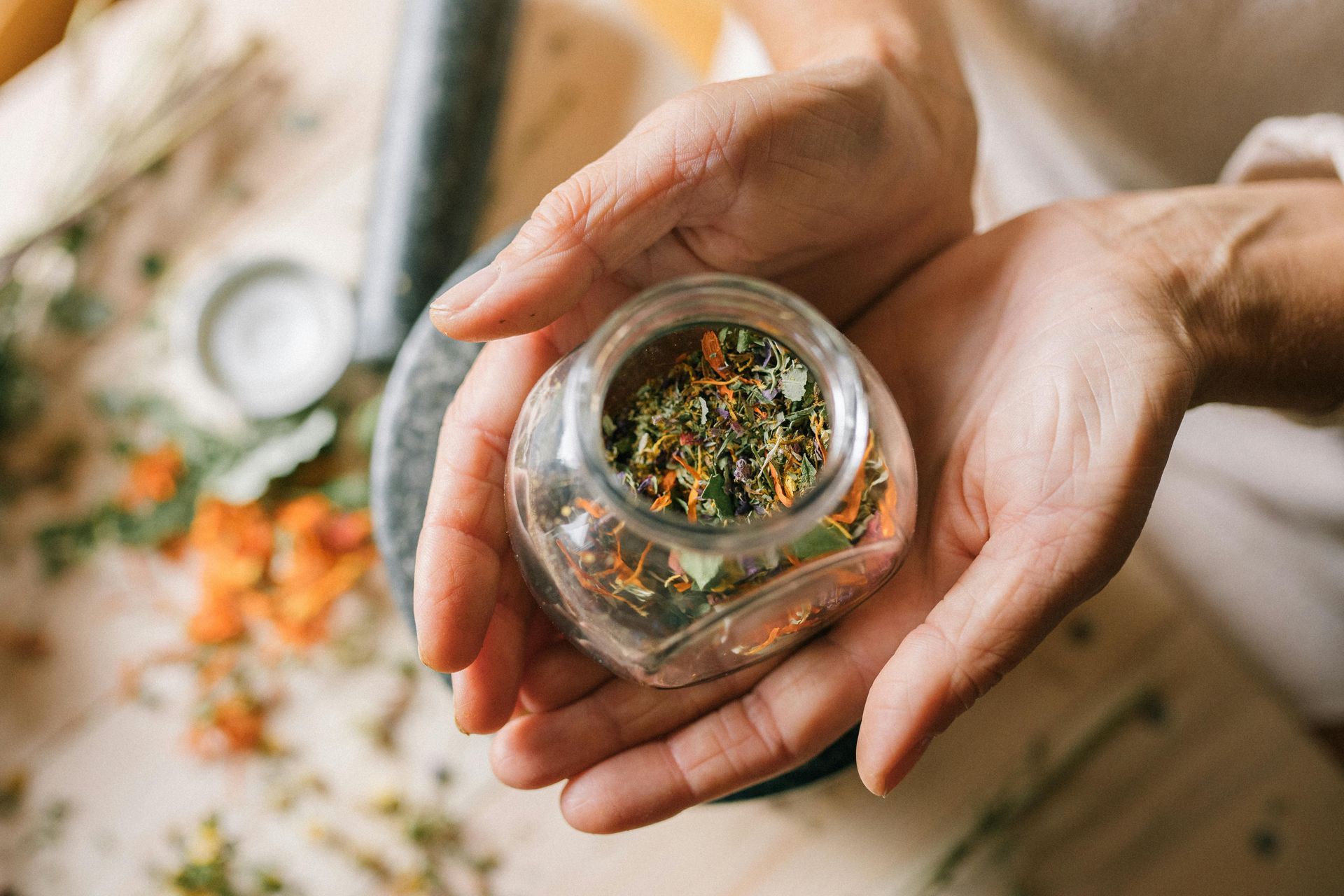 Hands holding a small glass jar filled with dried herbs, set among other herbs and mortar and pestle.