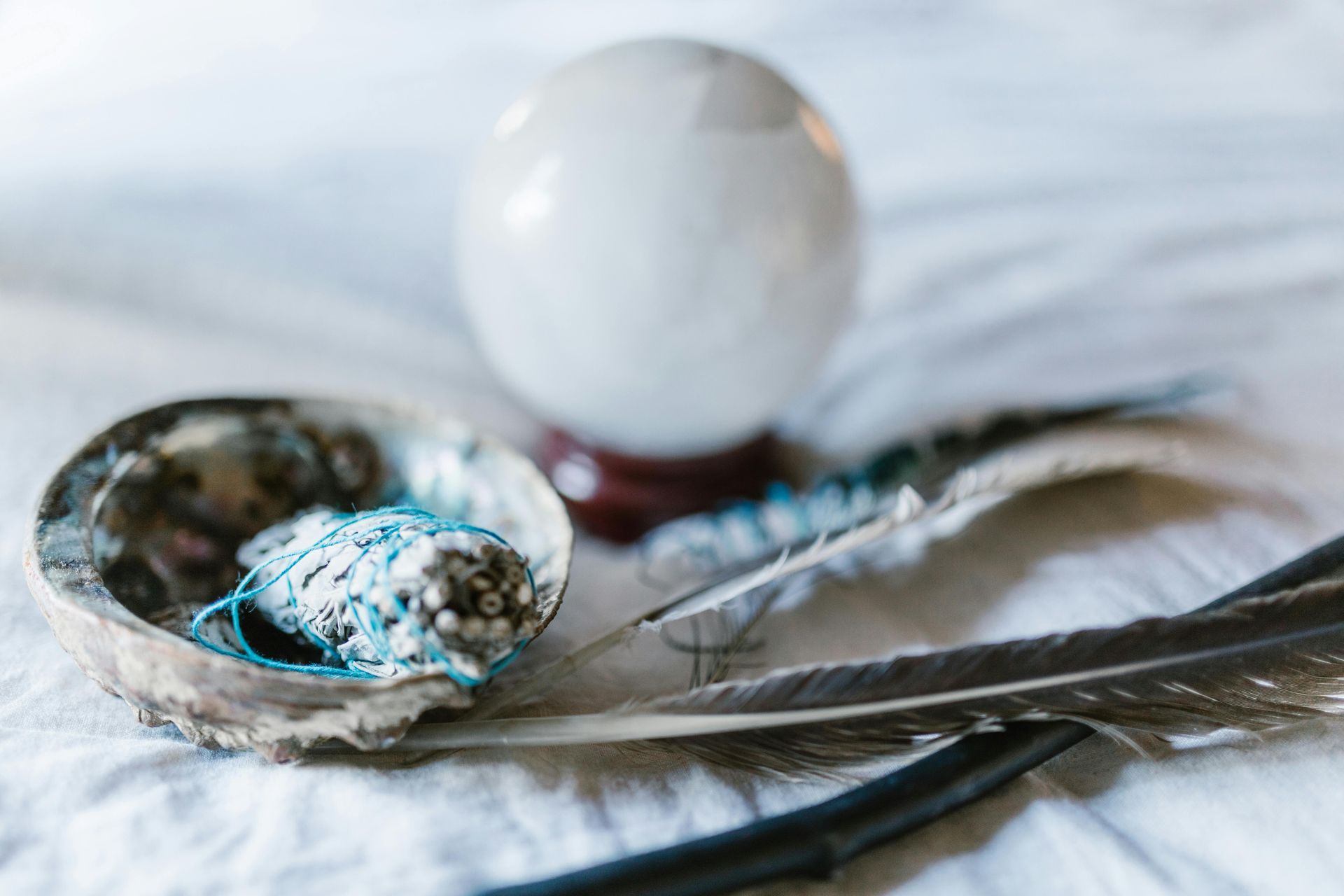 Crystal ball, feathers, abalone shell with sage on a white cloth.