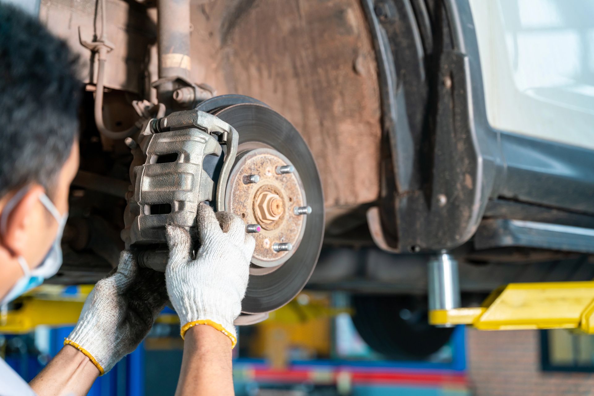 A mechanic in protective gloves inspects the brake assembly of a car raised on a hydraulic lift in a garage.