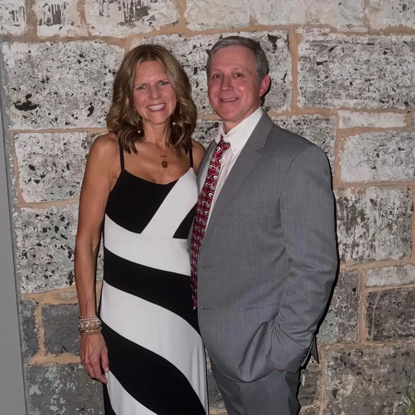 Woman in black and white dress and man in suit, smiling in front of a stone wall.
