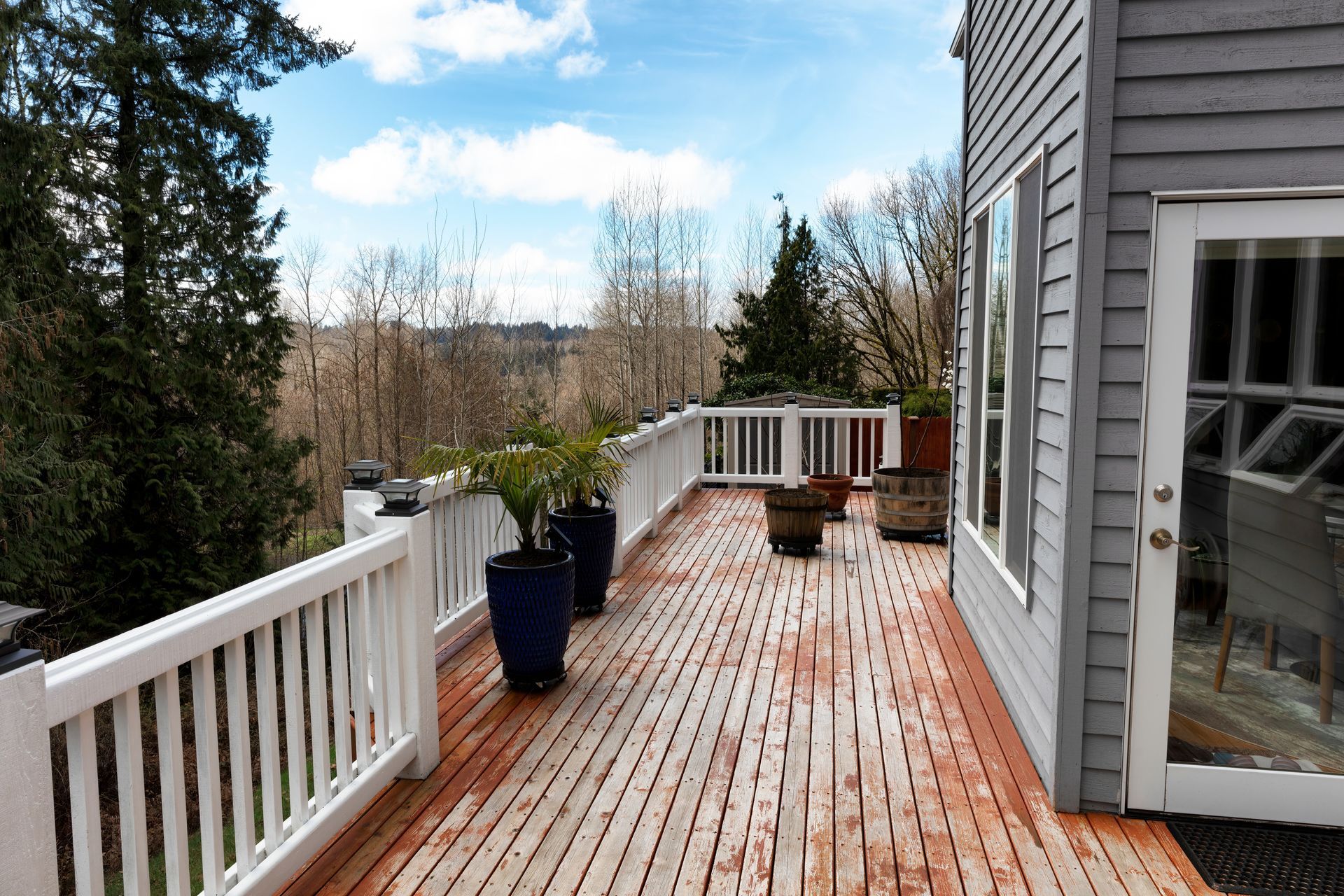 A long, wooden deck with white railings overlooks trees under a blue sky, beside the gray siding of a house.
