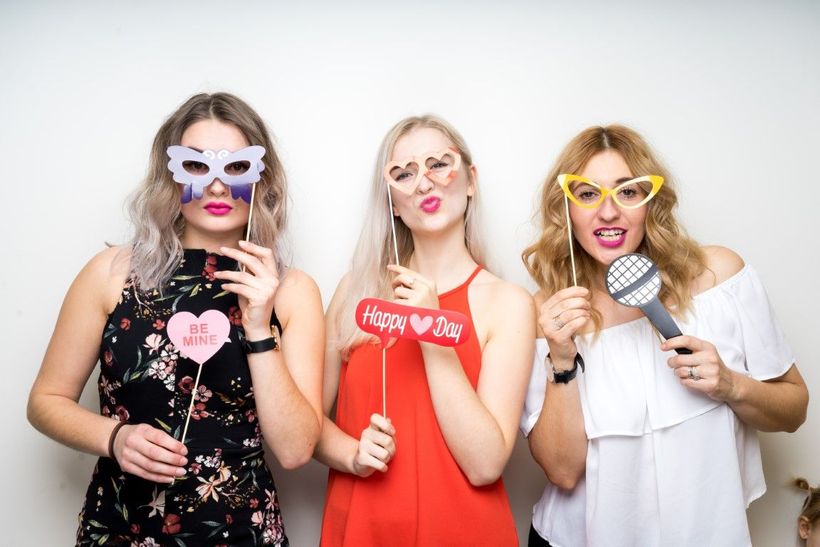 Three women posing in a photo booth with props; one wears a mask, another heart-shaped glasses, the third a microphone.