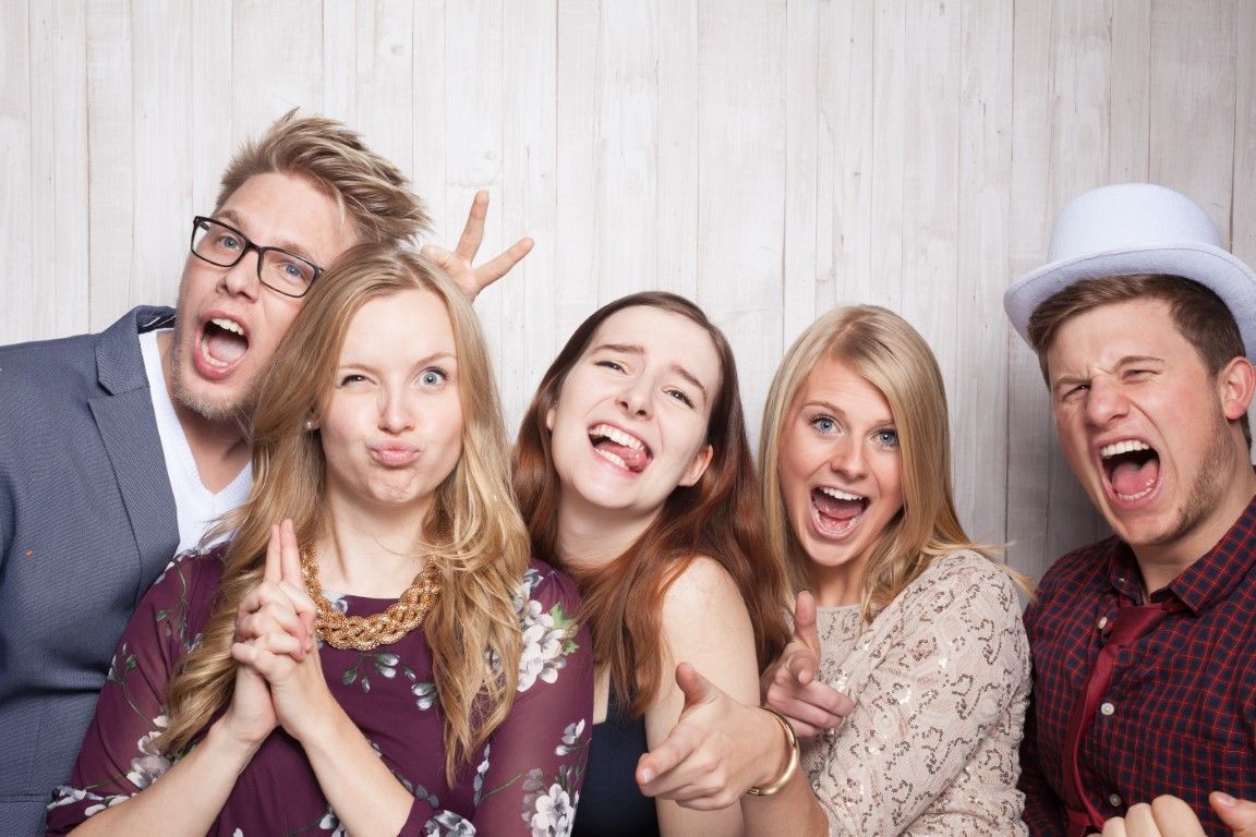 Five young people making silly faces in a photo booth; light wooden backdrop.
