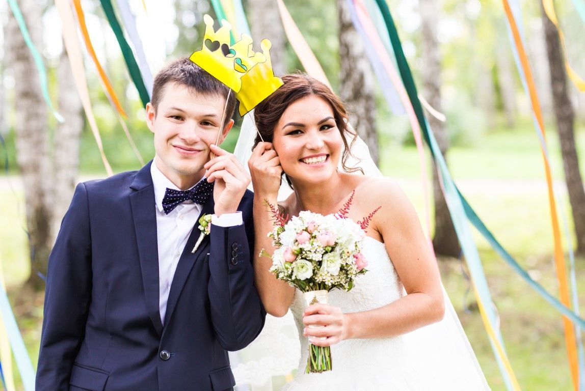 Bride and groom wearing paper crowns, smiling, outdoors with colorful ribbons.