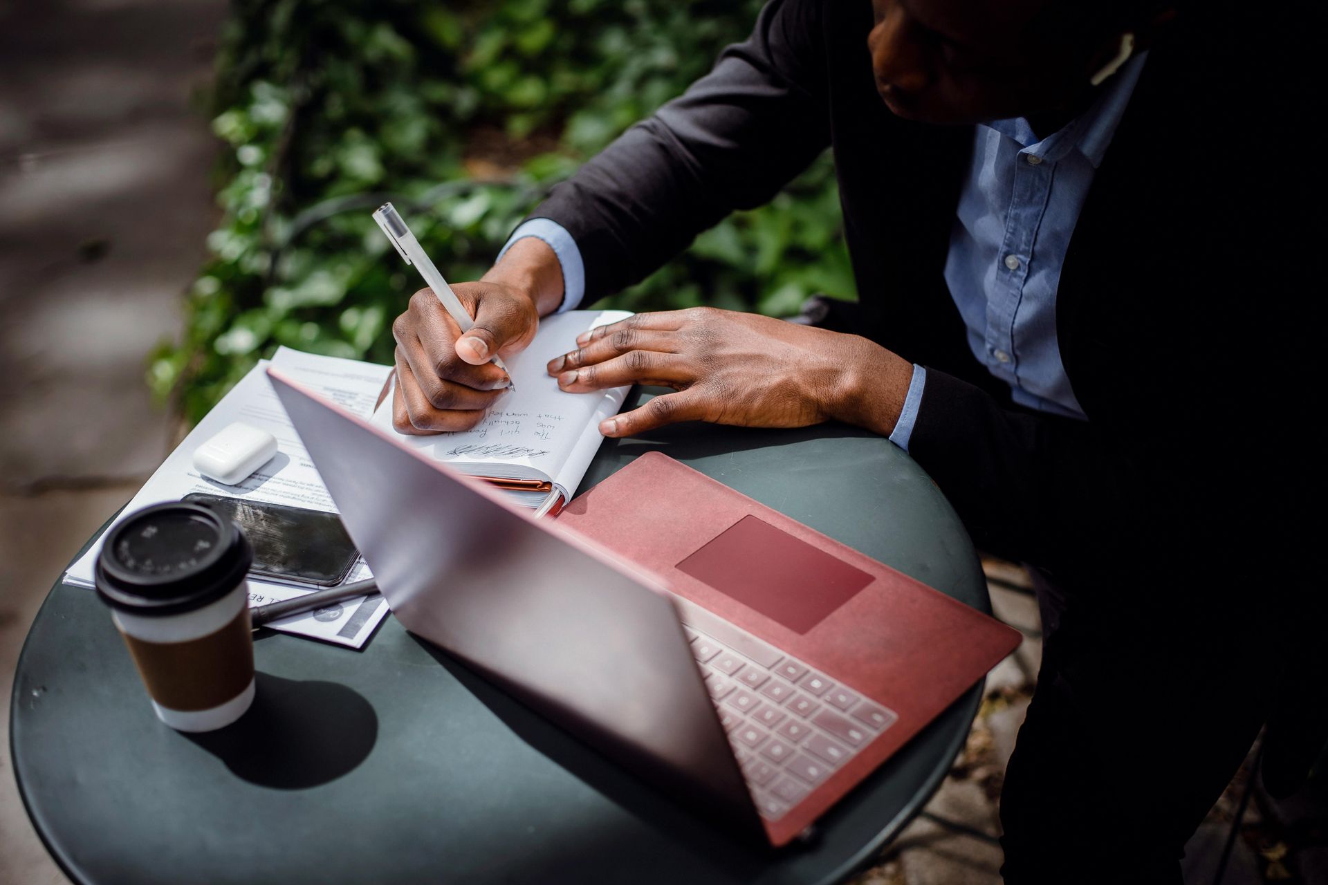 Person sitting at outdoor table looking at a computer and taking notes.  