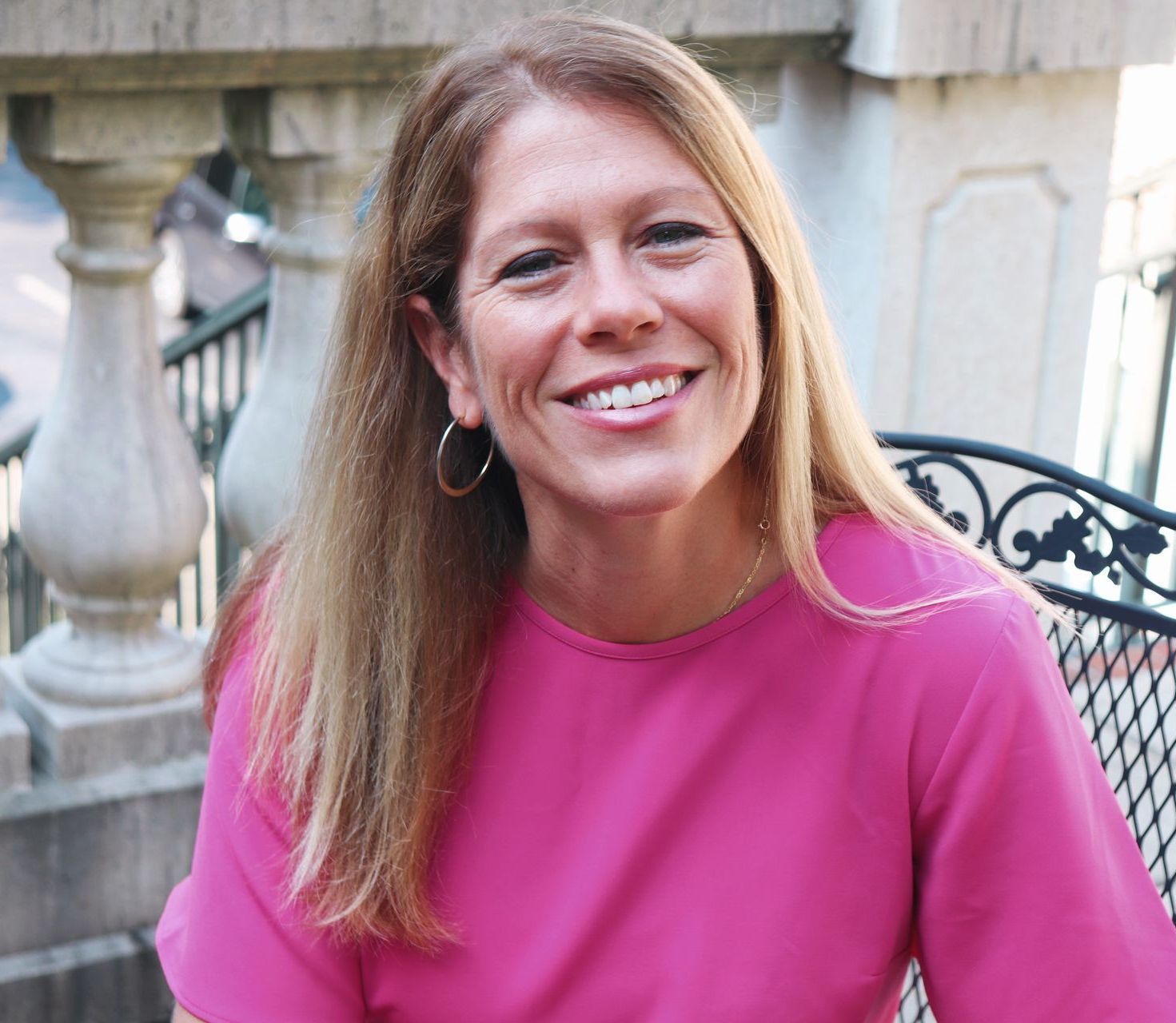 Woman with blonde hair smiling, wearing a pink top, seated outdoors.