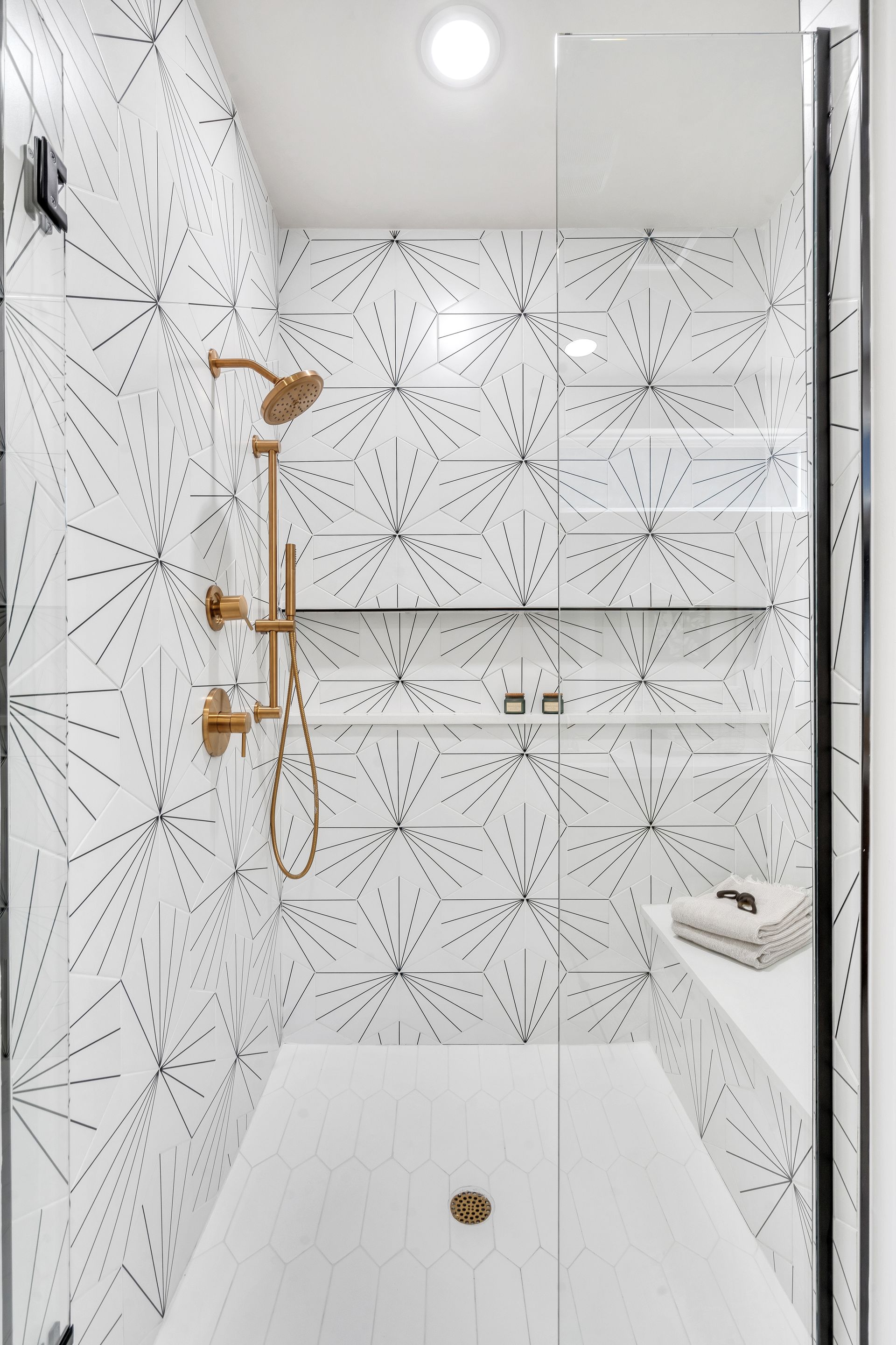 Small, modern shower with white and black patterned tile, gold fixtures, and glass door.