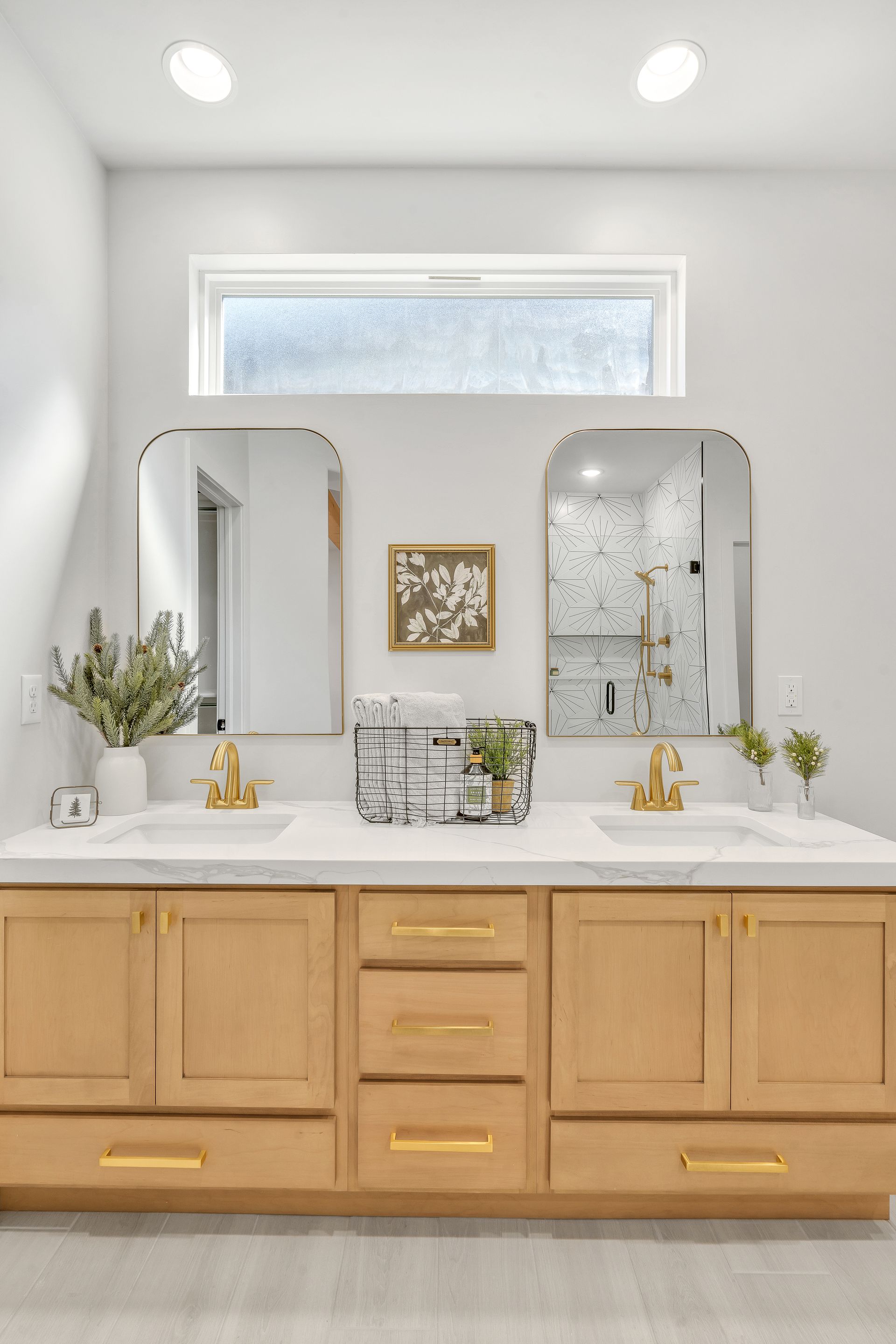 Bathroom with light wood double vanity, arched mirrors, gold fixtures, and a window.