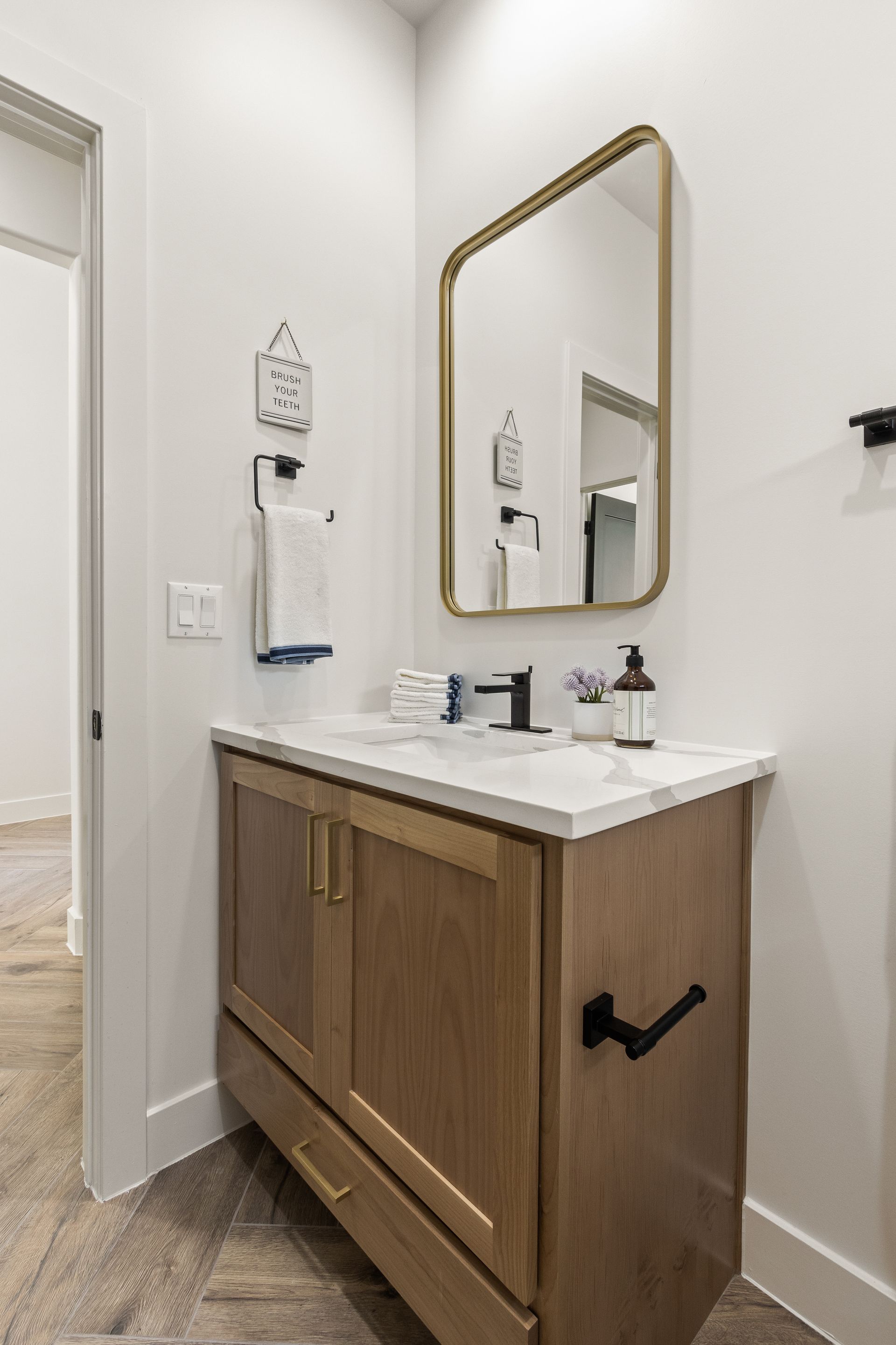 Bathroom with wooden vanity, gold mirror, white countertop, black fixtures, and white walls.