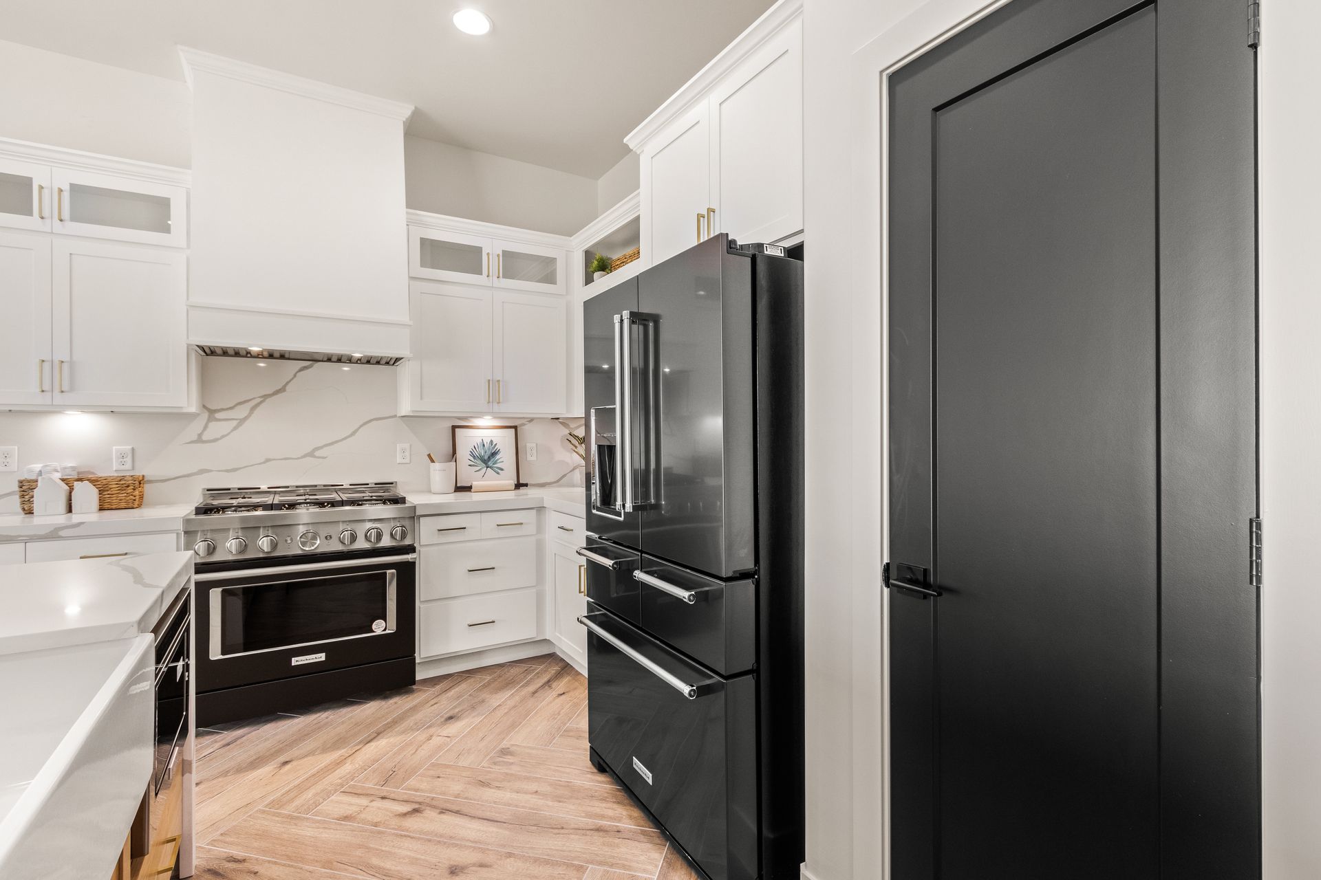 Modern kitchen with white cabinets, black refrigerator, and herringbone wood floor. Black door.