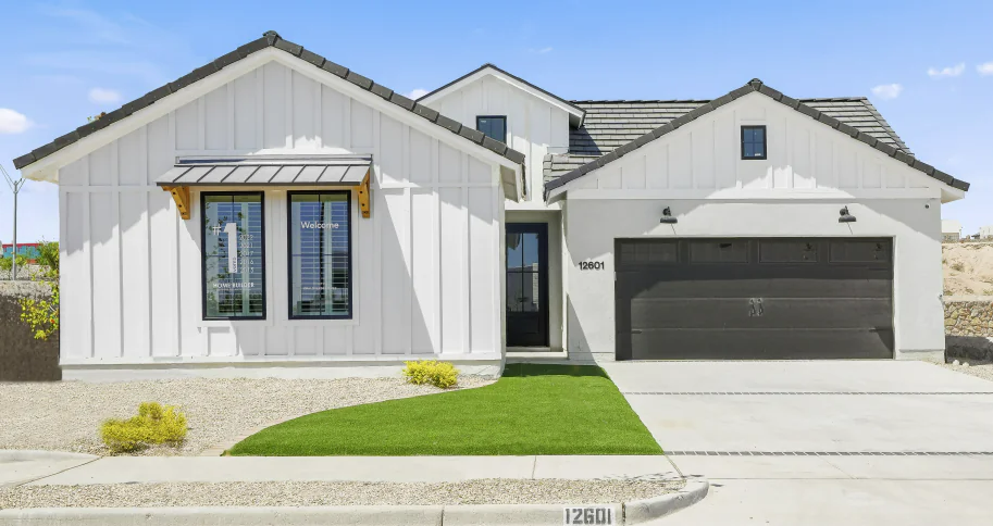 White farmhouse with black trim, garage door, and awning; blue sky, green grass, gravel yard.