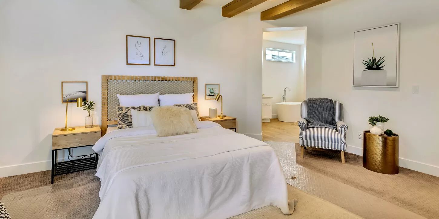 Bright bedroom with bed, nightstands, chair, and a bathroom entrance. White walls, wood beams, and neutral decor.