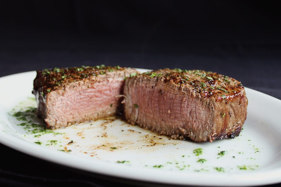 Sliced, cooked steak on a white plate with green herbs and a dark background.