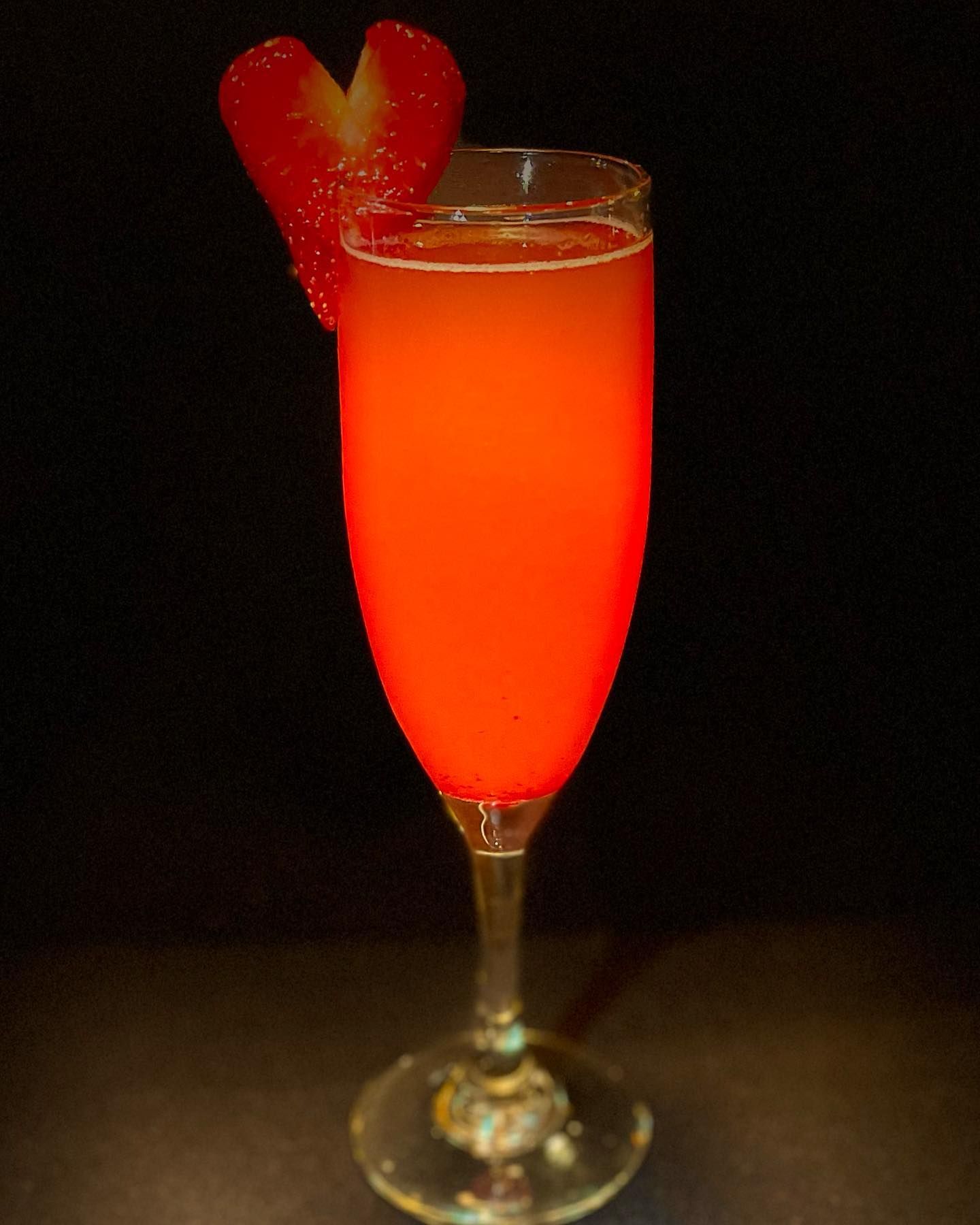 Red cocktail in a flute glass, garnished with a heart-shaped strawberry against a dark background.