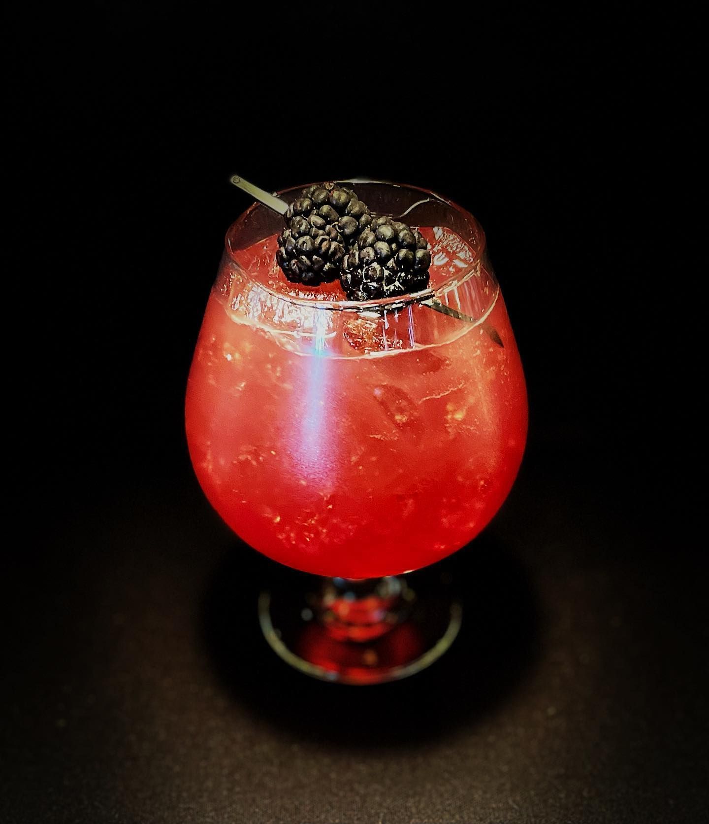 Red cocktail in a stemmed glass, garnished with blackberries, against a black background.