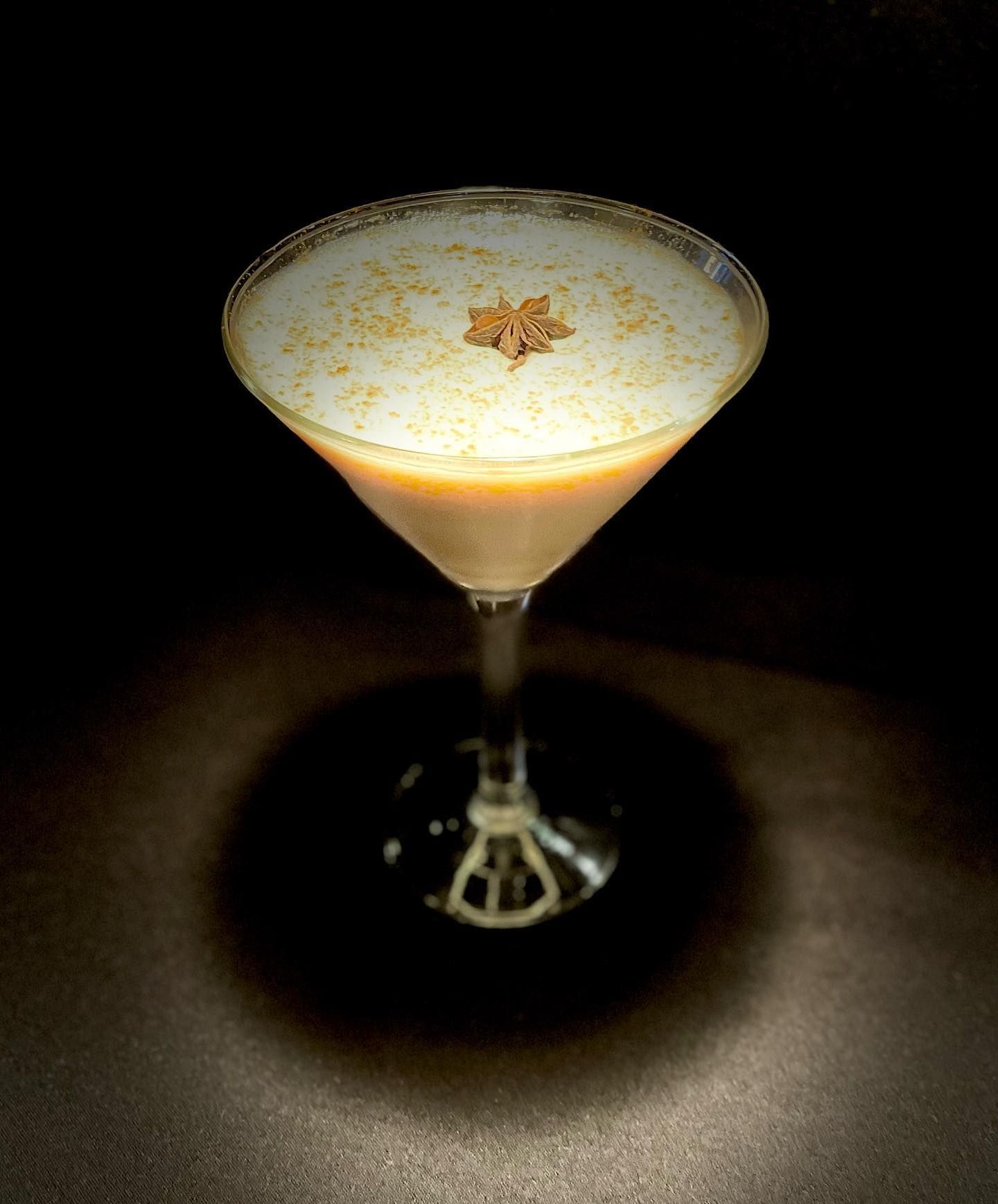 Cocktail in a martini glass, light brown liquid with foam, garnished with spices, dark background.