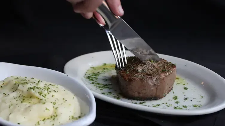 Person cutting a steak on a white plate, next to a bowl of mashed potatoes.