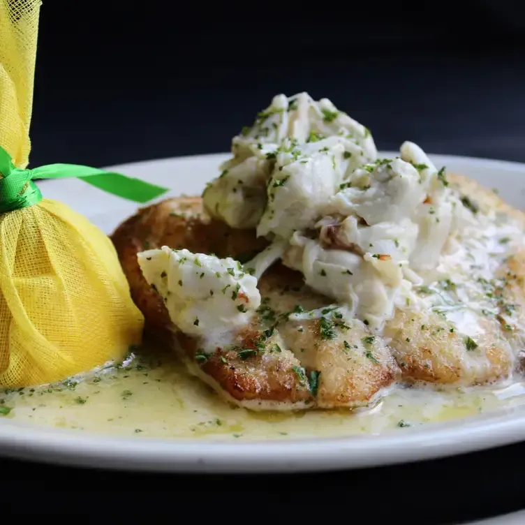 Pan-seared fish topped with crab meat and sauce, on a white plate with a lemon in a yellow pouch.