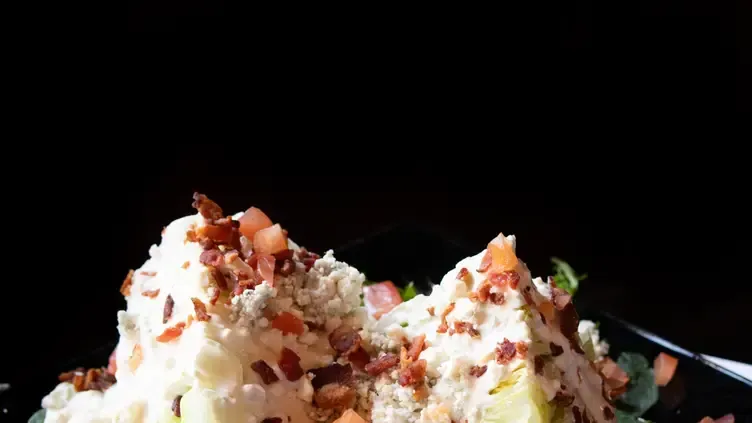 Wedge salad with blue cheese dressing, bacon crumbles, and a dark background.