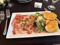 Beef carpaccio with capers, greens, toasted bread on a white rectangular plate. Black tablecloth.
