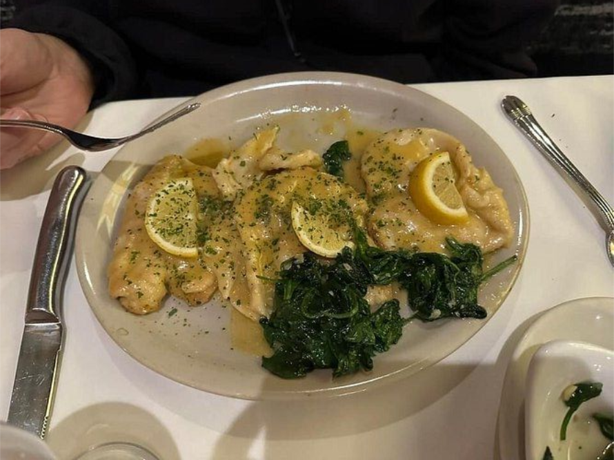 Plate of chicken with lemon slices, sauce, and spinach. Dining at a table with silverware.