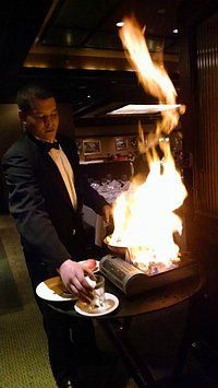 Chef in a tuxedo ignites a large flame while preparing food at a teppanyaki grill in a restaurant.