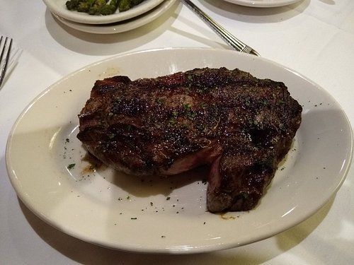Grilled steak on a white plate, with a side dish and silverware on a white tablecloth.