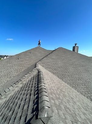 Person standing on a gray shingle roof under a blue sky, chimney visible.