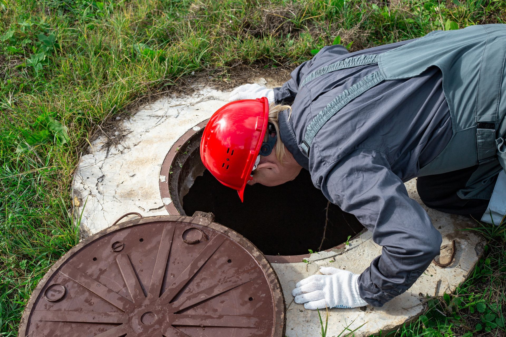 A man wearing a red safety cap and overalls peeks inside a residential sewer tank while crouching.