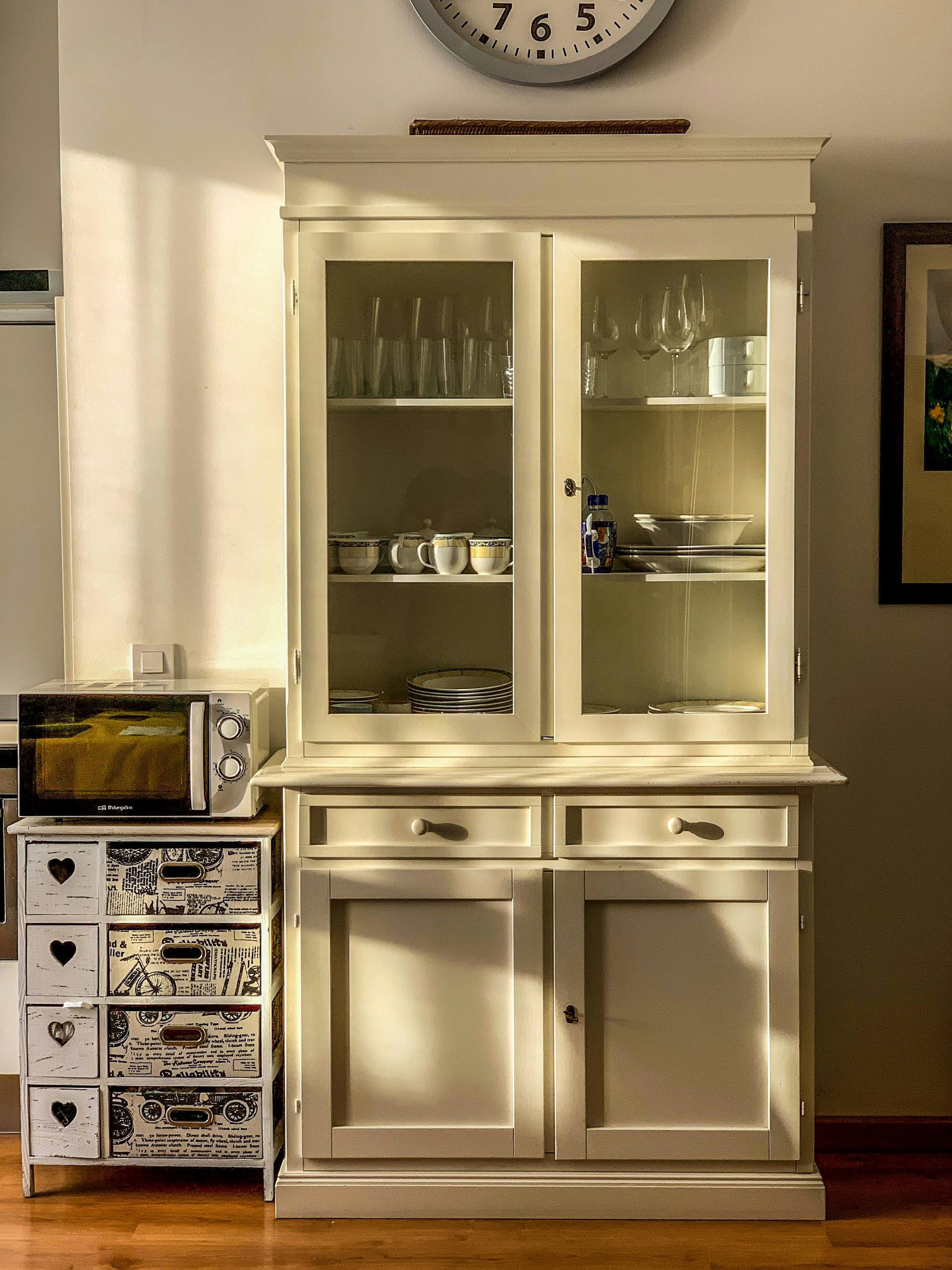 White hutch cabinet with glass doors and drawers beside a small storage tower in a sunlit room